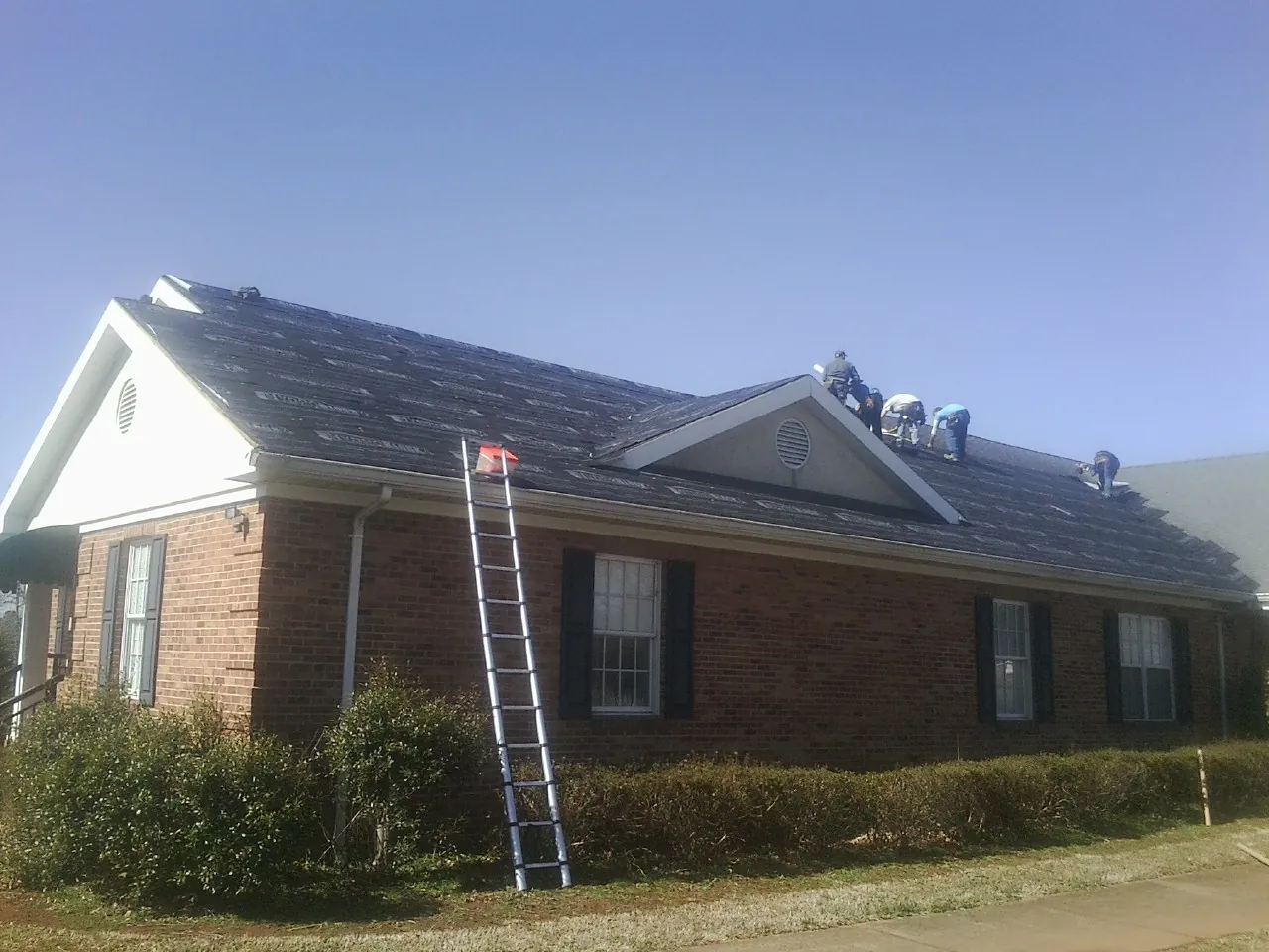 Crew tearing shingles off a brick ranch roof with a ladder set — Metal Roof Repair work by our crew in Darien Center