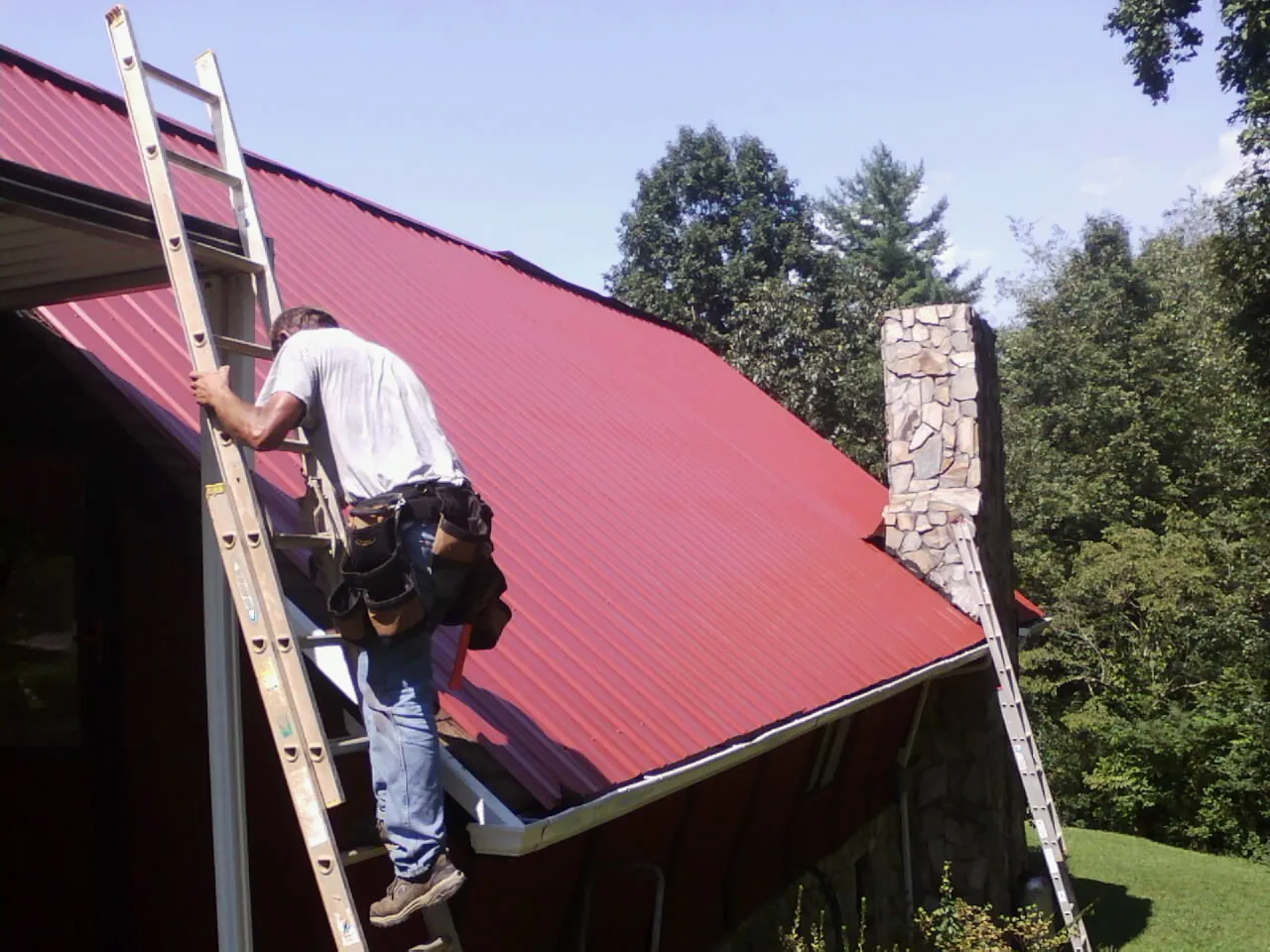 Roofer on a ladder beside a red metal roof and stone chimney — Soffit & Fascia Repair by Roofing Brothers in Darien Center
